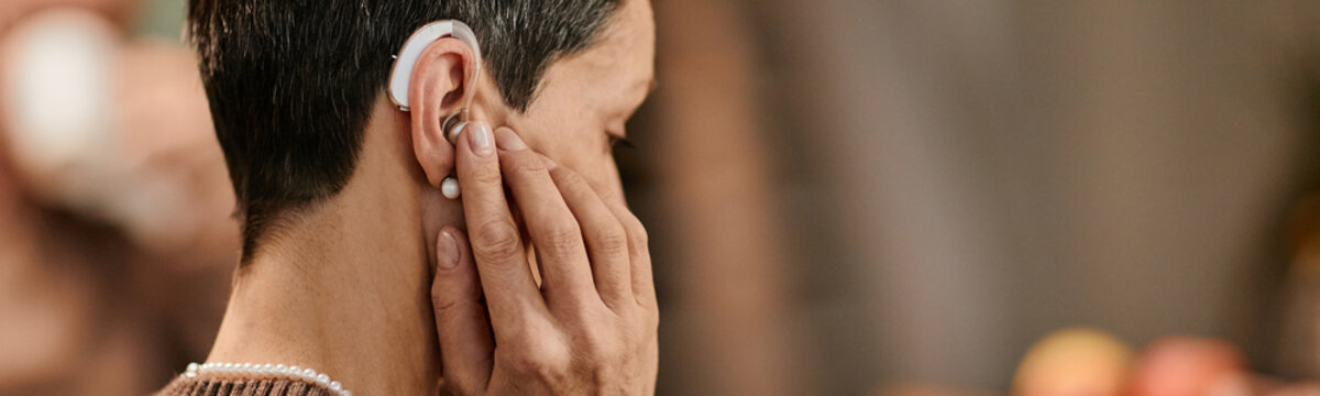 Website header shot of senior Caucasian woman adjusting hearing aid behind ear, short hair visible, hand gently touching device, indoor nursing home setting, focusing on assistive technology use