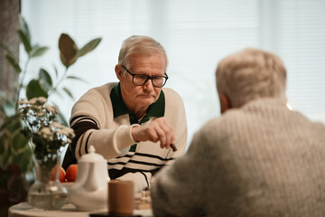 Senior Caucasian man playing chess with another senior man in nursing home, concentrating on making move, both sitting at table with chessboard, plants and teapot visible in background