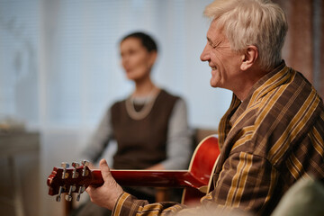 Senior Caucasian man playing acoustic guitar while smiling, sitting indoors with blurred middle...