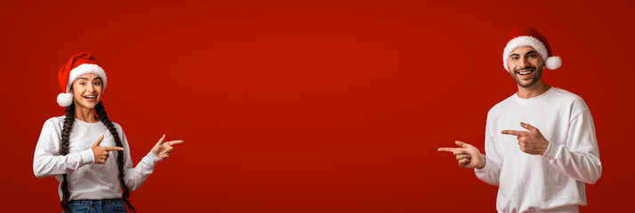 A joyful couple stands against a vibrant red backdrop, both wearing Santa hats. They point...