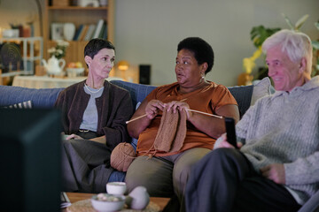 Senior Caucasian woman sitting beside senior Black woman knitting while senior Caucasian man holding remote control sitting on sofa in nursing home, watching television together