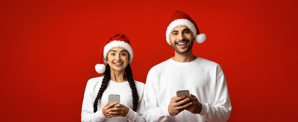 A joyful couple is wearing festive Christmas hats while holding their smartphones. They are smiling at the camera against a vibrant red backdrop.