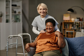 Obraz premium Caucasian middle aged woman caregiver standing behind seated Black senior woman smiling in nursing home setting, both women looking at camera, walker visible beside chair