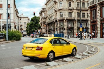 View of a vivid yellow taxi cab, symbol of urban mobility, navigates a European street framed by classic architecture and lush greenery, Budapest, Hungary.