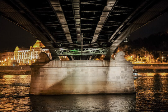 View of the bridge's sturdy stone base meets the shimmering waters, reflecting the city lights and dark metal of the bridge above, Budapest, Hungary.