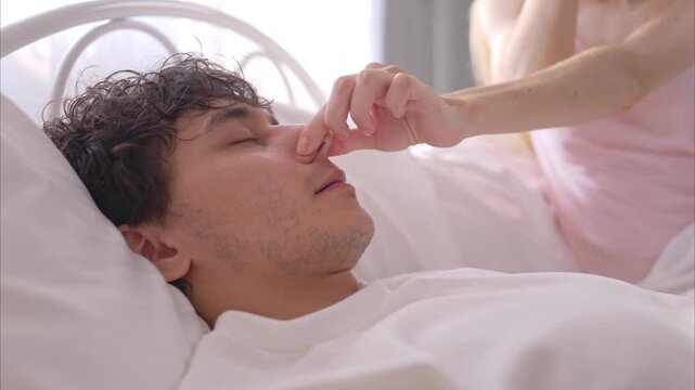A young woman covering mouth of her husband with hands while her partner snores loudly next to her in bed