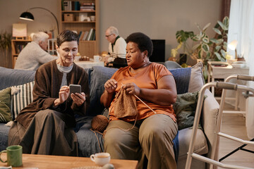 Obraz premium Senior Caucasian woman showing smartphone to senior Black woman knitting on sofa, background showing senior men interacting at table in nursing home, walker visible in foreground