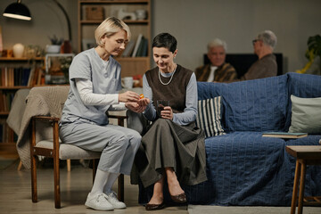 Caucasian female nurse assisting senior Caucasian woman with medication while sitting together on sofa in nursing home, two senior men conversing in background
