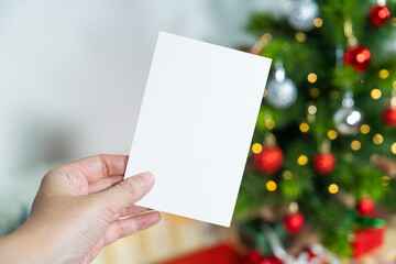 Hand holding a blank white card in front of a  blurred decorated Christmas tree with clipping path, perfect for holiday greeting design or mockup.