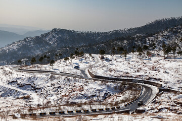 View of a winding road cutting through a snow-dusted mountain landscape under a soft sky, a tranquil winter scene of adventure and serenity, Murree, Punjab, Pakistan.