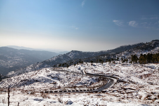 View of a winding road cutting through a snow-dusted landscape, flanked by dark green trees under a clear sky, creating a striking contrast, Murree, Punjab, Pakistan.