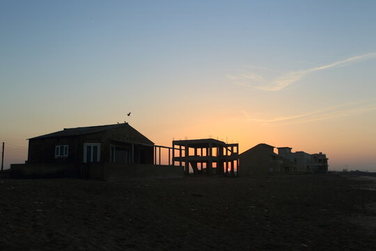 View of the sun setting behind silhouettes of buildings under construction and completed houses, casting a warm glow on the horizon, Sandspit Beach, Sindh, Pakistan.