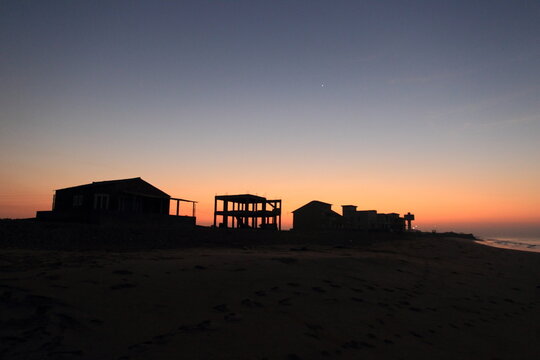 View of dark silhouettes of buildings and structures against a vibrant, gradient sunset sky over the tranquil beach, Sandspit Beach, Sindh, Pakistan.