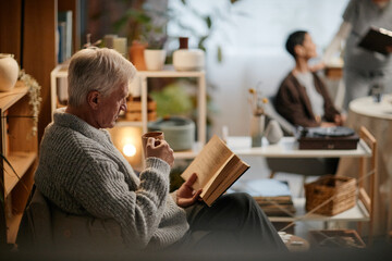Senior Caucasian man sitting in nursing home reading book and drinking from mug while relaxing, with blurred elderly woman and young adult caregiver interacting in background