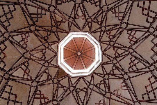 View of an elaborate ceiling with geometric patterns converging towards an octagonal skylight, casting a soft glow in Shahi Hammam, Lahore, Pakistan.