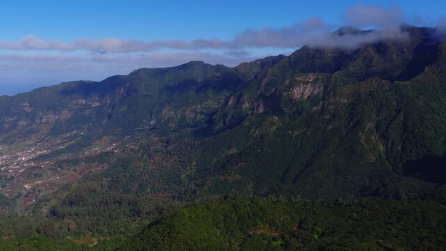 Drone moves over Madeira, Portugal mountains, steep green ridges, volcanic cliffs, laurel forest, cloud capped peaks, villages, Atlantic on horizon in bright daylight.