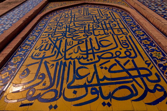 View of intricate blue calligraphy artfully adorns a sun-baked yellow wall, framed by ornate brickwork, creating a striking visual narrative, Lahore, Punjab, Pakistan.