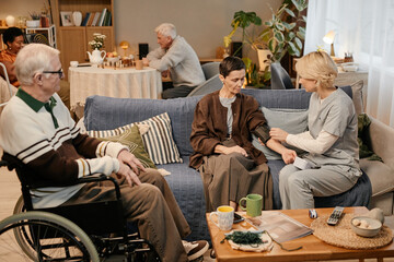 Senior Caucasian woman sitting on sofa receiving blood pressure check from young adult Caucasian female nurse, while senior Caucasian man with disability in wheelchair observing in nursing home
