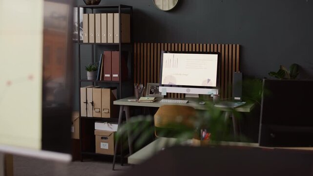 Computer screen showing presentation with diagrams at desk with office supplies and documents, black organized office shelf with plant in pot and paper folders placed near desk