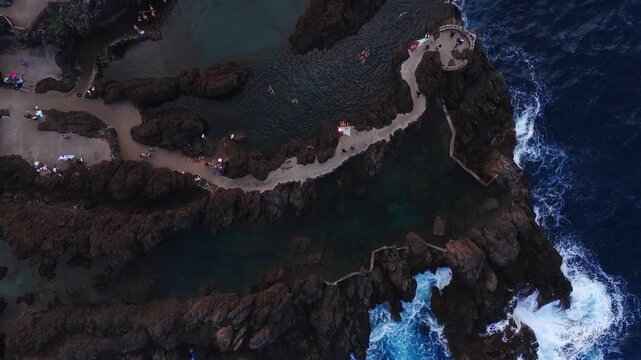Aerial top down view of Porto Moniz lava pools on Madeira, waves crash, swimmers float, visitors walk a paved path and circular platform in soft evening light.