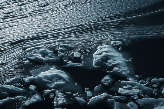 View of icy fragments scattered across the dark, rippling waters, creating a stark contrast between frozen stillness and flowing movement, Bedretto, Ticino, Switzerland.