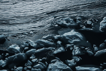 View of icy rocks clinging to the shimmering, rippled water's edge, a dance of frozen textures and fluid motion under a cold, stark light, Bedretto, Ticino, Switzerland.