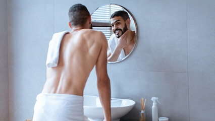 A man stands in front of a round mirror, wearing a towel, examining his reflection. The bathroom...