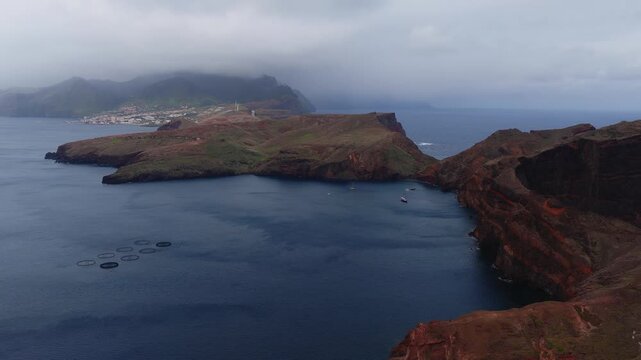 Aerial sweep shows rust red cliffs, walking paths, and a narrow peninsula on Madeira. Circular fish cages, anchored sailboats, and Canical under low cloud appear.