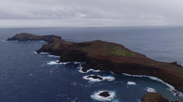 Aerial passes Ponta de Sao Lourenco, Madeira, Portugal, showing rust red soils, black cliffs, sea stacks, deep blue waves, and a distant lighthouse in overcast light.