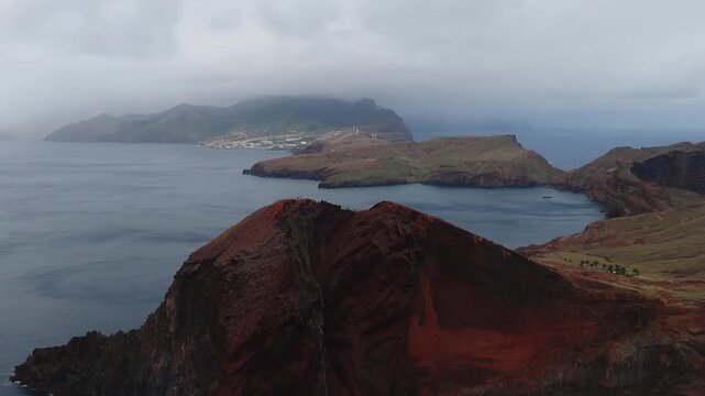 Aerial pan over rust red cliffs at Ponta de Sao Lourenco, Madeira, with Canical harbour, aquaculture pens, moored boats, and distant wind turbines under low clouds.
