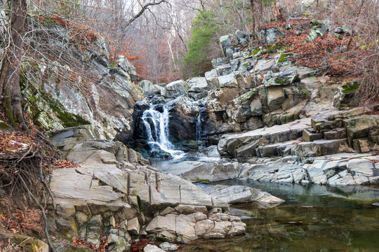 View of the cascading waterfall plunges down rugged, grey rocky cliffs into a serene pool surrounded by leafless trees, Scott's Run, Virginia, United States.