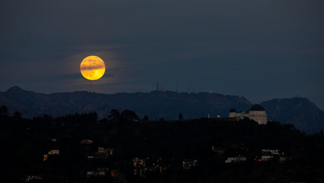 View of a bright, luminous full moon hanging over the silhouette of a mountain range, with the Griffith Observatory in the distance, Los Angeles, California, United States.
