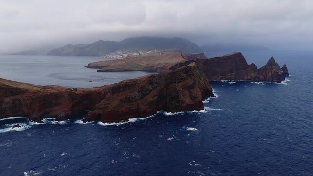 Aerial wide pan shows red brown volcanic cliffs, sea stacks, and a sea arch at Ponta de Sao Lourenco, Madeira, Portugal, with distant misty mountains and surf.