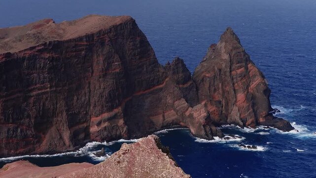 Aerial view shows red banded volcanic cliffs and a jagged sea stack at Ponta de Sao Lourenco, Madeira, Portugal, as waves churn below under daylight with high contrast.