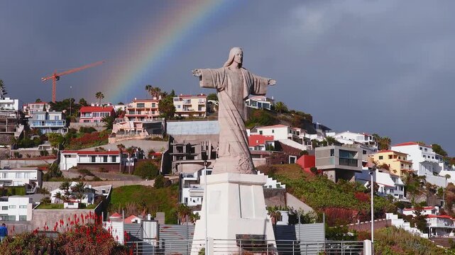 Aerial drone pans around Cristo Rei near Funchal, Madeira, as a rainbow arcs above steep terraces and cliffside homes in late afternoon light with deep gray clouds.