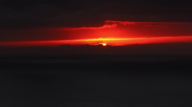 Cinematic aerial view shows the sun descending behind clouds over the Atlantic off Madeira, Portugal. Crimson orange light rims clouds and spreads across the horizon.