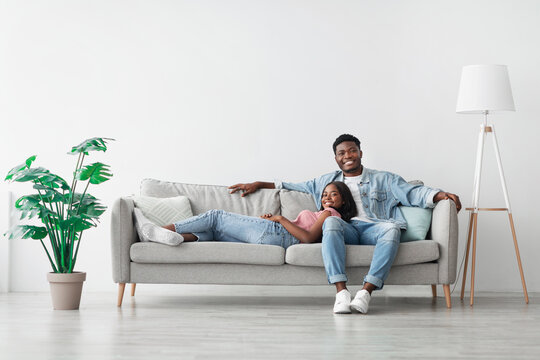 Weekend. Beautiful African American woman and handsome man relaxing on couch at home in living room, smiling couple looking at camera, posing, spending leisure time together, lady lying on guy's lap