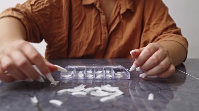 A woman with long white stiletto nails meticulously sorts through a selection of artificial nails from a clear container, preparing for a do-it-yourself manicure or nail application.