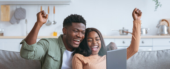 A joyful couple sits on a couch, raising their hands in celebration while watching a screen. Their expressions show happiness and excitement in a cozy living room setting.