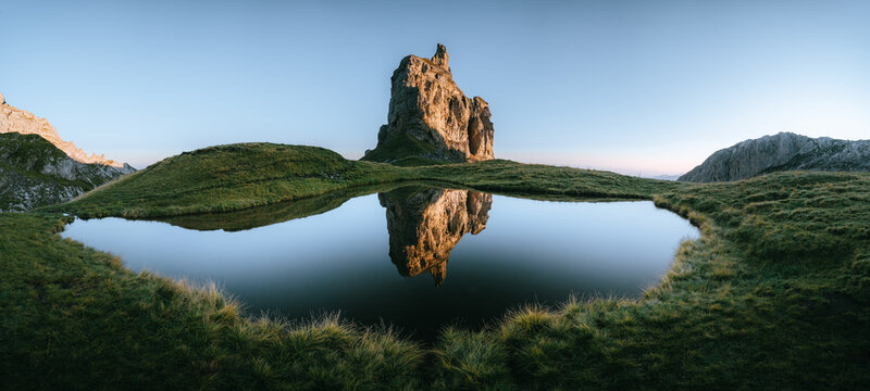 View of a monolithic rock formation mirrored on the surface of a tranquil lake surrounded by lush green grass under a clear blue sky, Muotathal, Schwyz, Switzerland.