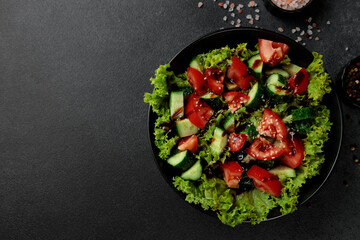 Vegetable salad of tomatoes, cucumbers, onions, lettuce dressed with aromatic dressing in a black plate on a gray background