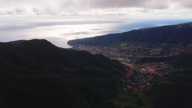 Aerial drone sweep shows a curved headland, red tiled roofs, and steep evergreen mountains near Funchal, Madeira, Portugal, with bright ocean and shadowed hills in daytime.
