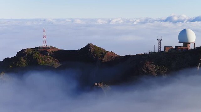 Aerial pan shows Pico do Arieiro radome, towers, cliffs, paths, and platforms on Madeira, Portugal as clouds fill valleys in soft early morning light with visitors visible.