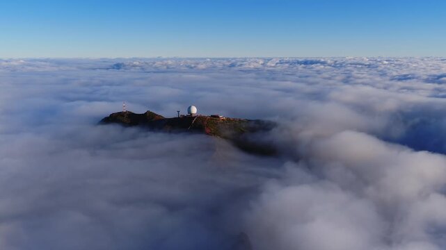 Aerial view shows a white radome and red and white antenna on Pico do Arieiro, Madeira, Portugal, as clouds roll around rugged cliffs in soft low angle light.