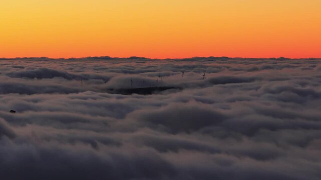 Turbine blades rotate above a moving cloud deck as a dark ridge rises on Madeira Island at sunset, with warm backlighting and strong silhouettes adding depth.