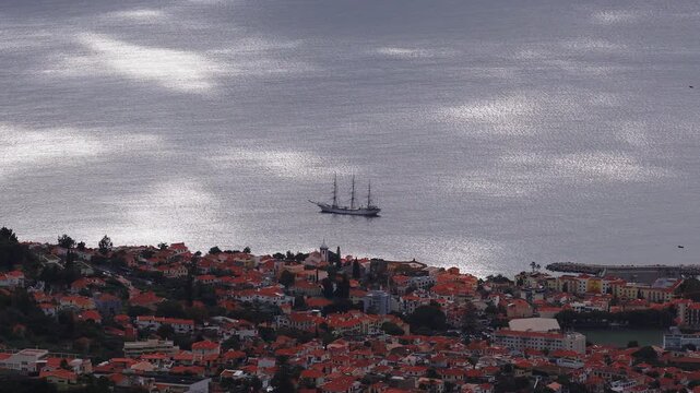 A high aerial view of Funchal, Madeira shows terracotta roofs, a church bell tower, a harbor breakwater, and a tall masted sailing ship gliding in late day light.