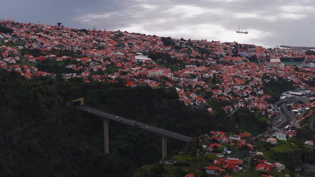 Aerial view of Funchal, Madeira, white houses with terracotta roofs on steep hills, a concrete viaduct to a tunnel, a harbour and three masted ship in late day light