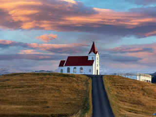 The red hilltop church stands boldly beneath a dramatic sky of red and pink hues, surrounded by lush green fields and snow-capped mountains on Iceland&rsquo;s Sn&aelig;fellsnes Peninsula.