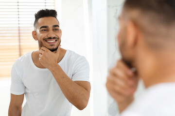 A young man is looking into a mirror, smiling and grooming himself. He appears relaxed and...