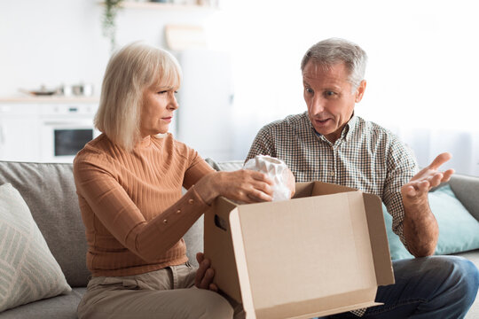 A couple sits on a couch in a bright living room, opening a box together. They appear engaged in conversation about what is inside the box, sharing expressions of curiosity.